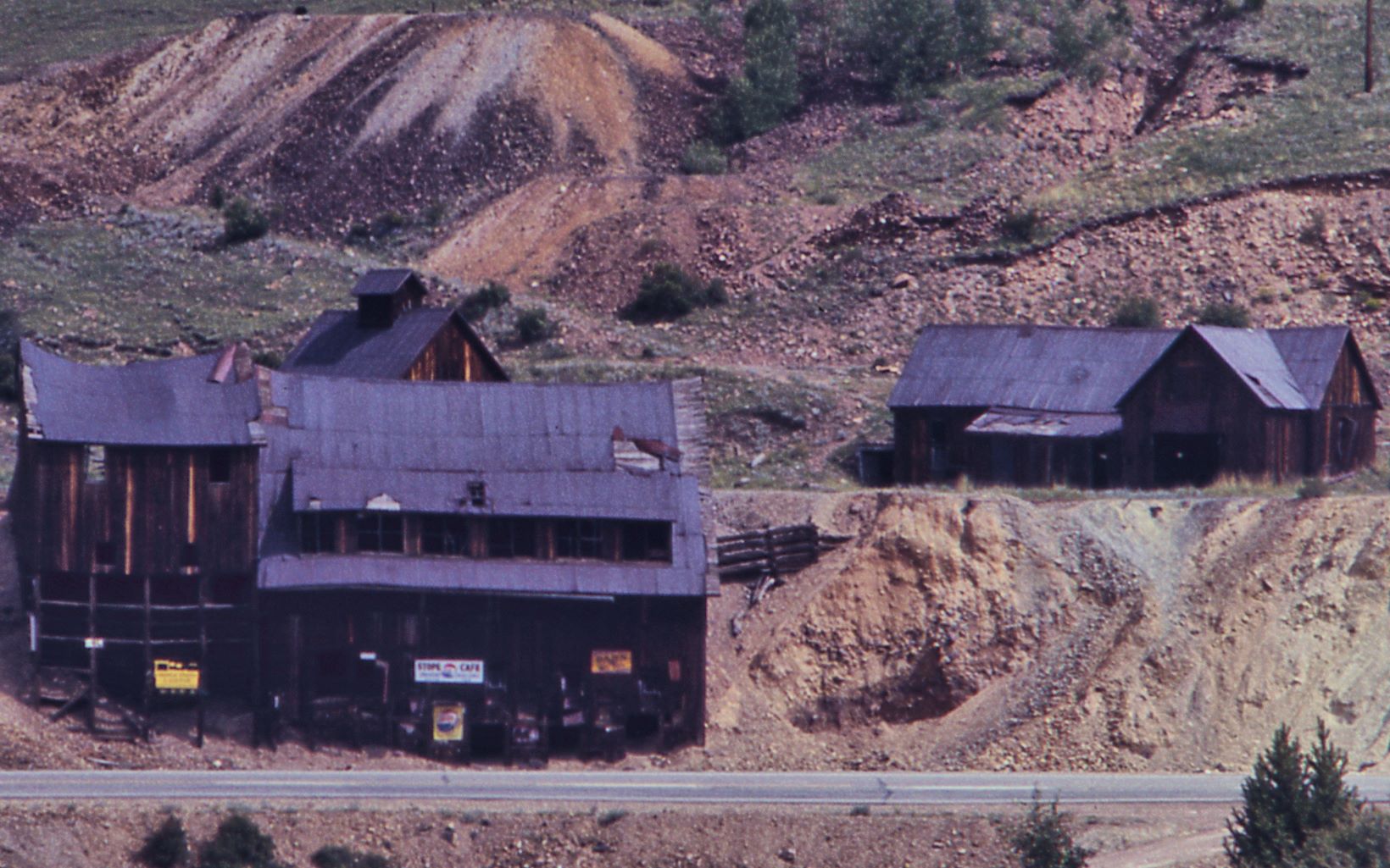 This cropped view of a slide photo from 1968 shows what is left of the Anaconda Mine structures. The Road in front of the Ore-House was the former F. & C.C. roadbed, which turned into a road when that railroad was gone, and which was later the main road between Victor and Cripple Creek towns, but today all this is gone, road and everything.
   Closest to the road is the Anaconda mine Orehouse, with the Blacksmith and its cupola visible behind it, and to the right is what appears to be a tunnel opening, just left of what appears to possible be the Powerplant Structure when looking at a Sanborn 1908 Fire Insurance Map, or the Office, maybe also just be a shed like structure. Behind the structures is the former roadbed of the Midland Terminal seen, at about middle top/down, coming from left-hand side, and entering a cut behind the shed/possible powerplant structure, at the right-hand side.