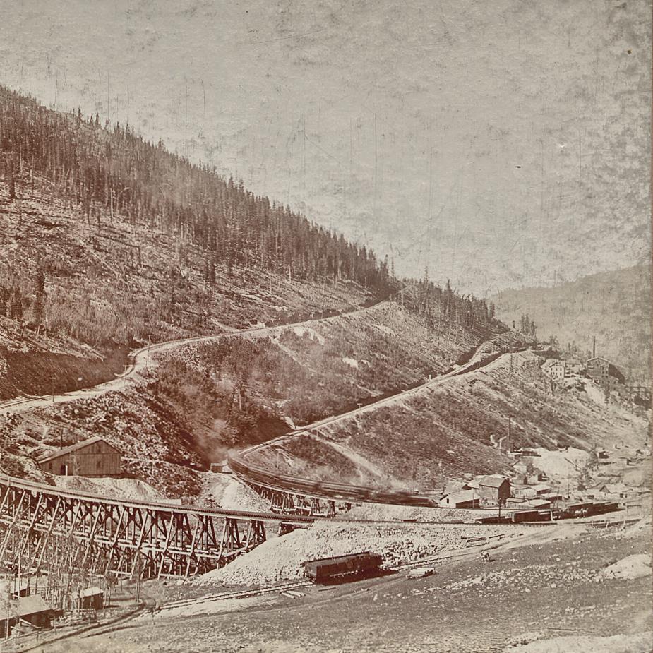 This view was taken just above the M.T. trestle crossing Eclipse Gulch, and it shows an early look at quite a large shaft house of the Eclipse Mine, and how it is located between the roadbeds of the M.T on the upper side and the F. & C.C. on the lower side. Photographed around 1899/1900, as we see the Economic Mill way down the Eclipse gulch at the right-hand side, making this 1899 or later as that is the year the mill was built. And as I see no traces of the Low Line which was built in 1900, this was photographed in that timeframe.
