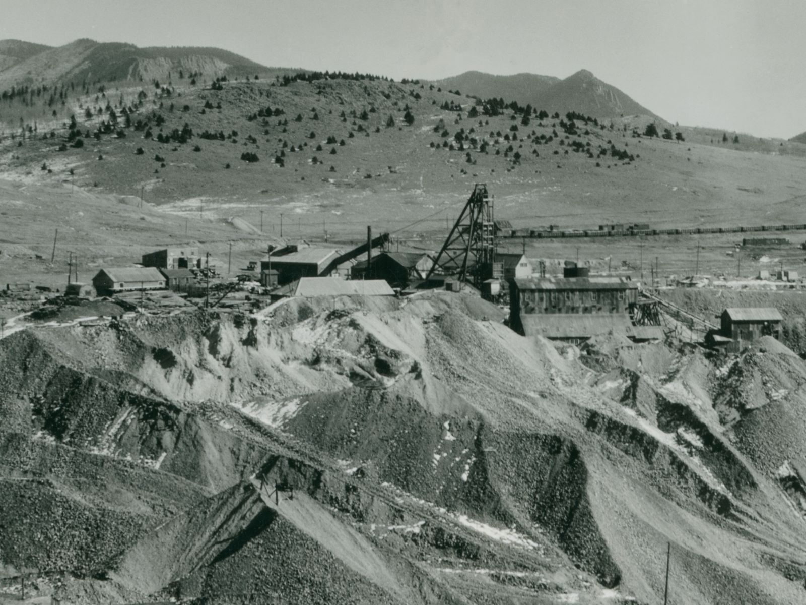Cropped view of the Vindicator Mine Operations 1934-ish style. Huge dumps covering the ground, the Ore-House is partly buried in them it appears. Not the best view, but it is a long distance shot from near the Last Dollar mine on bull Hill, so hard to get a good quality shot.
   Behind the large Headframe is seen a huge string of boxcars in the Bull Hill Yards of the Midland Terminal, making up the background scene behind the more top part of the headframe of the Vindicator, and of course the mountains in the horizon background.