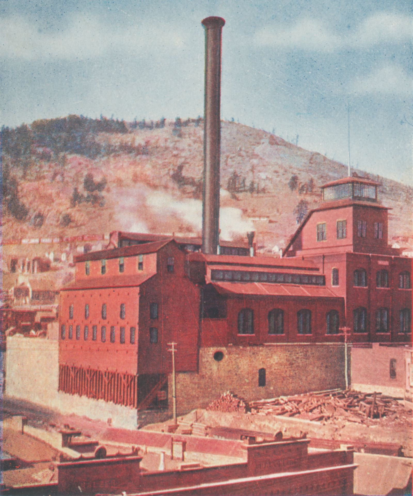 This view shows the Gold Coin Mine in Victor in all its glory, focusing more on the Ore-house. One get to see the foundation walls which still for the most part is still there in Victor, with its brick based shaft-house with the stained-glass windows and tower like part over the top of the head frame. The smokestack is very visible in this view, and in the background, is the Squaw Mountain seen, with the mainline of the M.T. seen just behind the top of the orehouse.