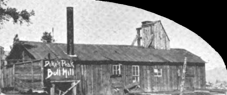 A view at the early shaft house of the Pike's Peak mine, it is a long narrow wood structure with a small cupola at one end and a smoke stack and external water tank at the other end. Also, a Shed is poking out towards the photographer at the left-side of this view, easy seen on the 1896 Sanborn Fire Insurance map of this mine. View is looking north.
On the DPL photo, there is more view to the left, and in front of this structure, I would advise to check that view out if interested in this mine here.