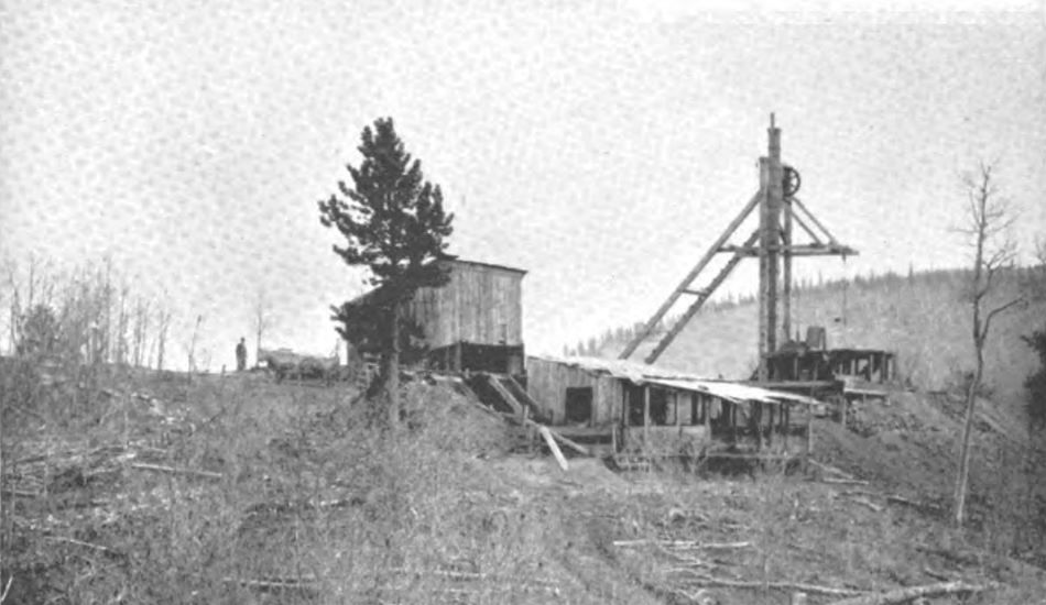 View at a small hoist house and a smaller head frame with a platform where the shaft is, at top of a small hill. With a single slope roofed shed and open shed somewhat closer to the photographer. Town of Victor would be located over the hill towards left.
