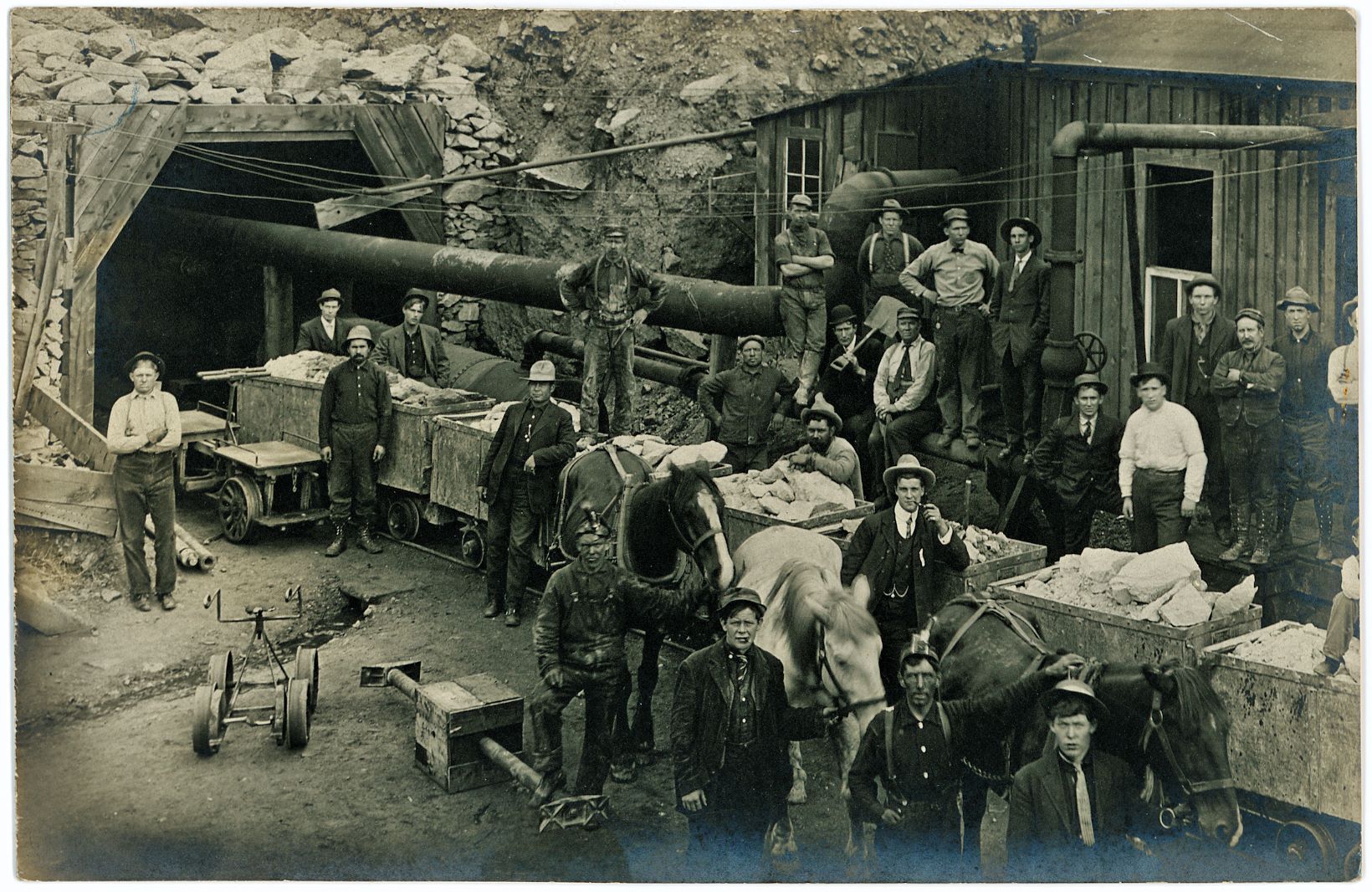 This Group-Photo of workers, management and equipment at the Portal of the Roosevelt Drainage Tunnel during its construction has been used in an article in the July 1914 edition of the Mining Science journal/magazine, so it predates that date with an unknown time. No names given in neither of my sources for this image, but there are many men here.
   Image shows 8-ore-cars filled with rock from the tunnel work, also three horses are held in front of the cars, uncertain if they where the motive power used to haul the cars out of the tunnel. There are also some other foot powered rail cycle types shown, a shed makes up some of the background, a compressor building of some sort as there is a large pipe coming from it into the tunnel opening.