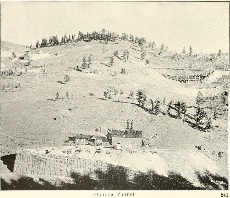 This view of lower Gold Hill is interesting in several ways as it shows several mines and three railroad lines.
* At bottom front is the Ophelia Tunnel seen, also later known as Moffat tunnel, or also known as the Gold Exploration tunnel, with its large crib wall and big power plant and tunnel house.
* Above the Ophelia is seen first the roadbed of the 3-foot narrow gauge F. & C.C with a trestle at left-hand side and a fill near right hand-side where a trestle used to be.
* Further up the hill then come the standard gauge M.T. railroad, with a trestle near right-hand side, which was later filled in and is today known as the ''Million Dollar Fill.''
* Near upper left is the roadbed of the Low Line, it enters into a large cutting somewhere left of the Pointer mine, sadly I am unable to pick out that cut and it has puzzled me for years why I can't see it.
* Near upper left is also the Pointer mine structures, and by looking at the PPLD view at 100% one can actually read the name Pointer on the ore-house of this mine and Pointer Mine on their shaft house!
* Just uphill, behind the Pointer, is another mine, they appear to have a head frame in front of their hoist house, I think that would be the Keystone Mine, but I am not certain. It could be a shaft on the Independence claim as that actually fits better with a claim map and a shaft on that, but I still think this mine would been known as Keystone.
* Left of the Pointer mine, up top at edge of view, there is the mine I know as Index Mine, but I also seen it as the Mint Mine before the Index mine came along, and possible this might been known as Keystone, Irish Molly or Goddard in early days as I think it is on one of those claims. This is before they built the large ore-house along the side-spur of the Short Line, branching off from the Low Line behind the hill so to speak.
* Also left of the Pointer, further down from the Index, is what I believe a structure on the Red Spruce, a small shed like, possible a hoist house, on the upper of two small dumps just above the M.T. roadbed.
* Just right off the Pointer mine is the structures of the Maggie No. 2 mine, the dump of the Pointer almost seems to engulf the small triangular head frame they seem to have on their property.
* Almost middle of view sideways, uphill from the Ophelia tunnel structure, there is a mine structure between the F. & C.C. and M.T. mainlines, quite a big head frame, hoist house and shed, water-tank and smokestack so more than a prospect type of mine operation. But I have a hard time placing it on a claim map and find a name for it, as it could on both the Puzzle & Bird claims. Possible others crossing same area, but for now I think it is the Puzzle mine structures.
—> There are more structures, but I am not able to decode things better at the moment.
   I did procure the colored version of this image, if that is what you see. Source was gray-toned, or in common speech black & white. Used an online service and tweaked and worked with image to get what looks best to my eyes at the moment.