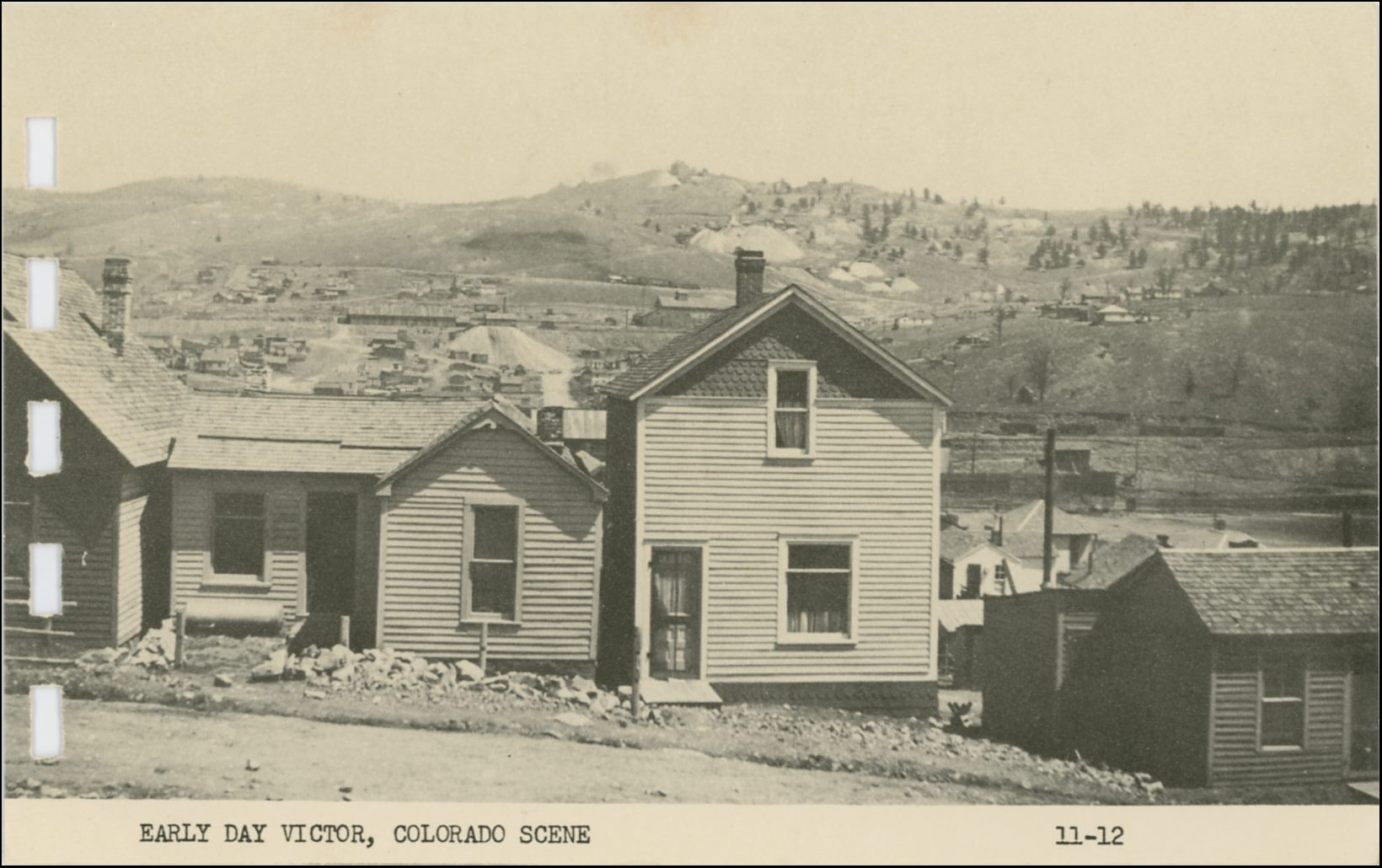 This view of Gold Hill from a street in southern Cripple Creek is mislabeled to be an early Victor scene for some unknown reason. Being this is from a printed card in a small more modern postcard folder it is not the greatest quality, but one gets an idea of the area it pictured. I've been unable to pinpoint the street in the foreground, but I think it might be the First Street, possible south Colorado Ave., but I doubt it as this don't look that far up the hill.
* Either way, about center left/right and 1/3 down from top, behind the two-story house in foreground, on get to see the north half of the Cripple Creek Sampler, located up along the M.T. roadbed, with the Short Line railroad below it, having come around the hill at right hand side. Incidentally, the original locating of the sampler was along the F. & C.C. grade seen just below the top/down center on the right-hand side, and would have been hidden by the same two-story house.
* About 1/3 down from top, and 1/3 from left-hand side, a long structure will be seen, that is the Trolley Barn for the High/Low Lines of the Electric System of the Short Line railroad. The big dump just right of the Trolley barn I think is part of the Cripple Creek & Gold Hill Deep Mining & Development Company operations.
* Straight up from the chimney on the before mention 2-story house there is a big dump that I think is part of the Midget operations.
* About center left/right and not fully 1/6 down from top, above the before mention Midget, is the characteristic shape of the large Shaft-House of the Anchoria-Leland mine seen against the sky at top of Gold Hill.