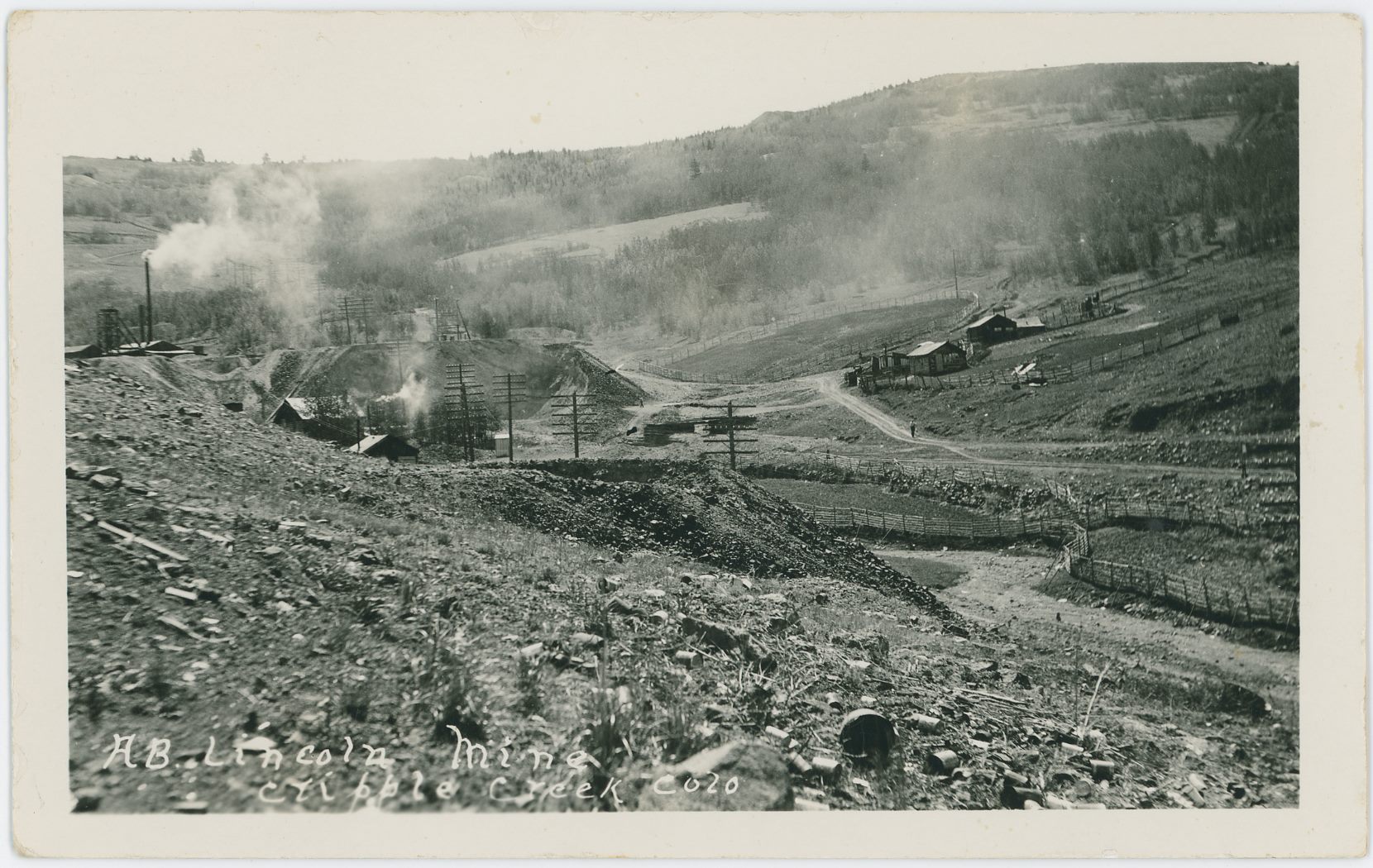 While this card is not marked, the Text Title has same type of writing as another card with the Young name on it, so I assume it is taken by the photographer named Young.
The Scene is in lower Poverty Gulch, looking towards Gold Hill in the background, with the large dump and operations of the Abe Lincoln Mine seen at the edge of the left-hand side, going into the image towards right. The Dump with a hole in the foreground I can't say at this moment in time [13.06.2017] which claim it was located on, so sorry I can't help with that.
What I can say is that I assume this view to be in the 1930's or so, and there are seen two switch-stands about middle top/down on the right-half-part of this view, where I think the one closest to the right-hand edge is the one for a siding at the M.T. Wye below the old Midland Sampler site, while the one to the left is the one for the upper spur that went to the top side of the same Sampler. There also appears to have possible been a loading dock of woods at the end of the spur near the end of the mine dumps of the Abe Lincoln.