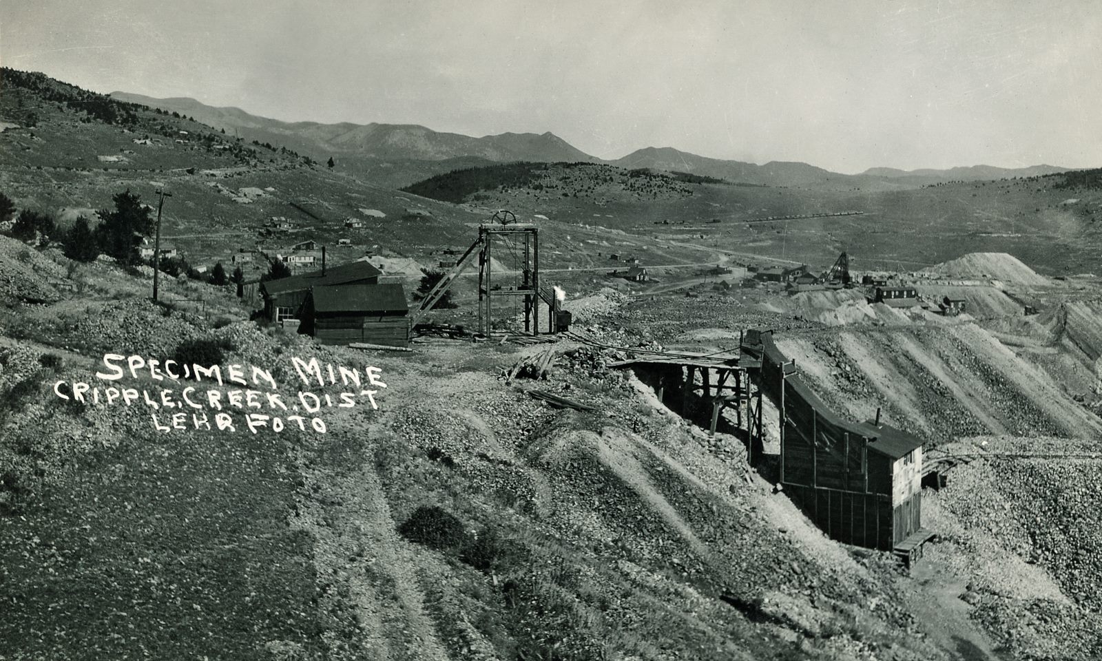 This view is a straightened, enhanced and cropped version of the 300dpi scan I did of the source postcard, showing the Specimen Mine Surface structures in the foreground. Direction of this view is east, northeast, on Bull Hill. The Specimen Mine operation is seen with an open Head-Frame and Hoist-house and shed linked to it, and a small Ore-house to the right down the hillside a small bit.
   In the distance, up from the orehouse, the surface structures of the Vindicator No. 1 Mine are seen with a large Head-frame and Ore-house down into massive dumps, impossible to tell if it still is linked to a railroad or not in this view. Further on in the distance to the left, a big string of railroad cars say that the Midland Terminal is still in operation, but there is no trace of the High Line Electric Division, as where that should be I only see a road surface – cutting almost the headframe of the Specimen Mine in two so to speak.
   Image is not sharp enough to tell if there are any rails left on the old Golden Circle tracks seen climbing Bull Hill, or is it Bull Cliff, about 1/4 down from top and about 1/4 in from left-hand side, but I do think the standard gauge M.T. tracks are there still when this photo was taken by Lehr many years ago.